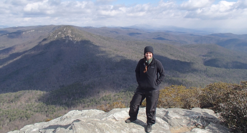 A person smiles while standing on an overlook, high above the tree-covered mountains below. 